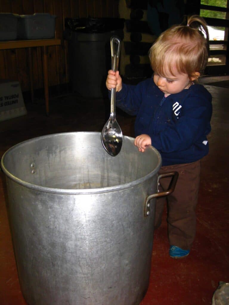 A toddler holds a giant spoon and pretends to stir a giant pot. 