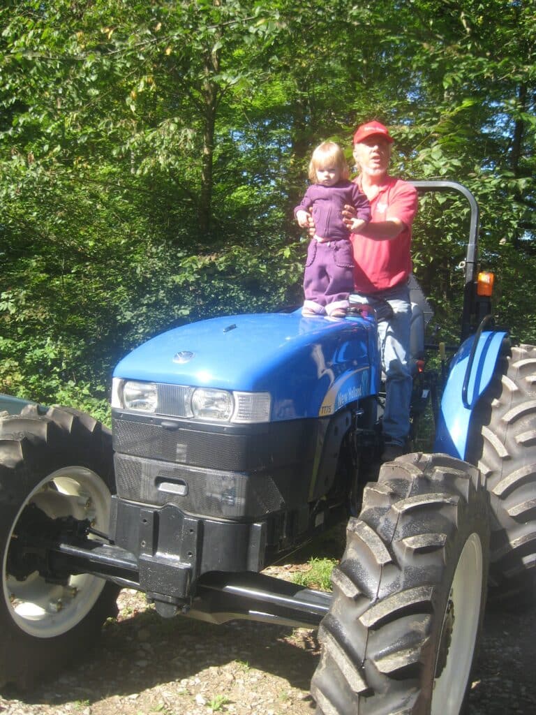 Dave, a man, stands on a tractor holding a toddler upright in front of him