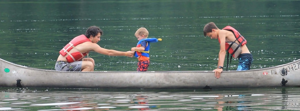 A toddler is standing in a canoe with two older campers