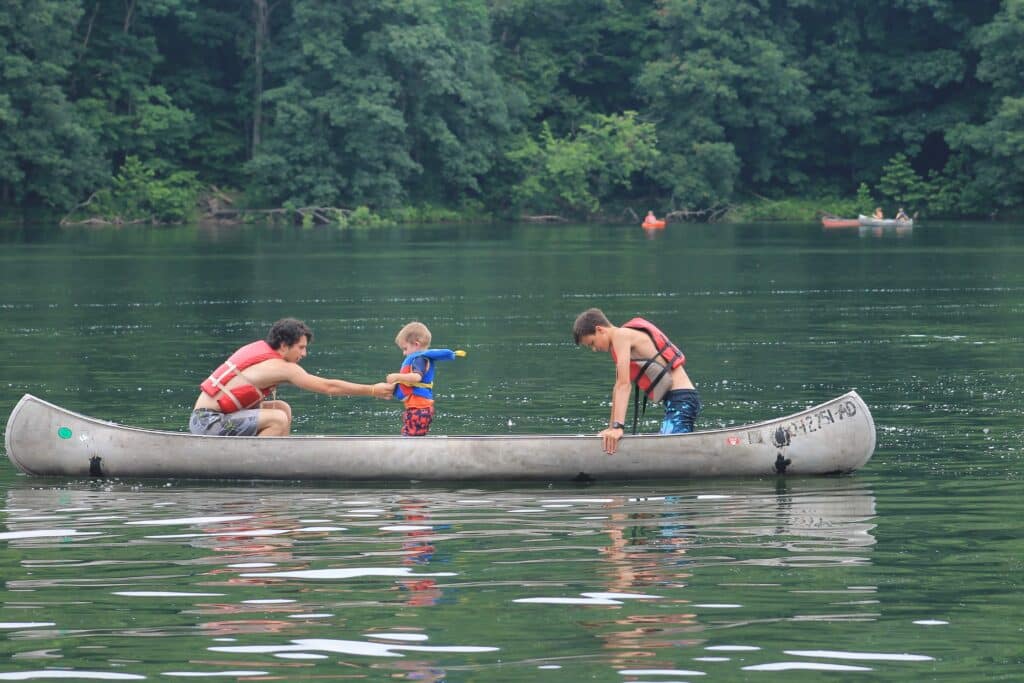 A toddler stands in the middle of a canoe with two older campers on either side. One adjusts his life jacket. 